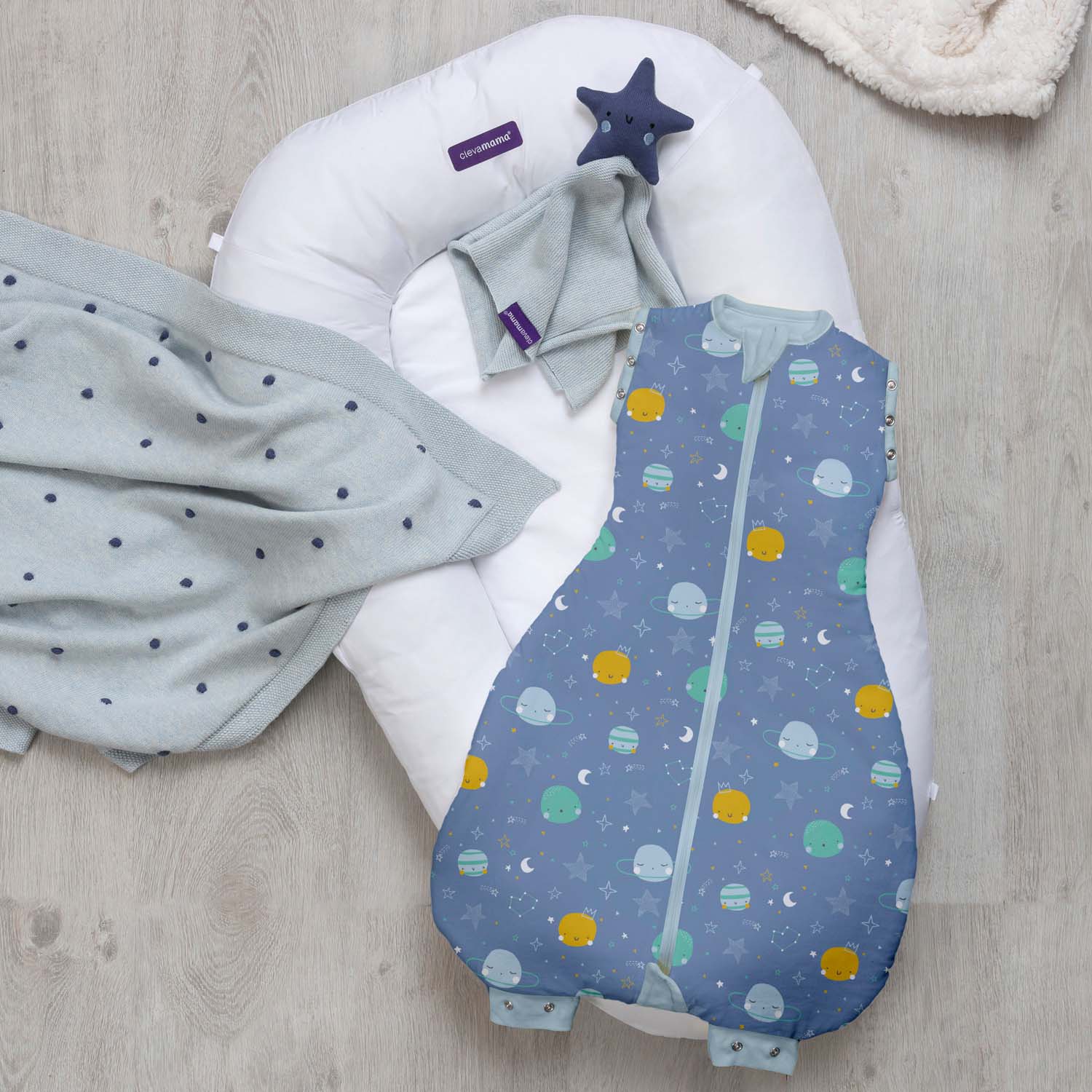 Baby sleeping bag with space design, gray swaddle, and white pillow on a wooden surface