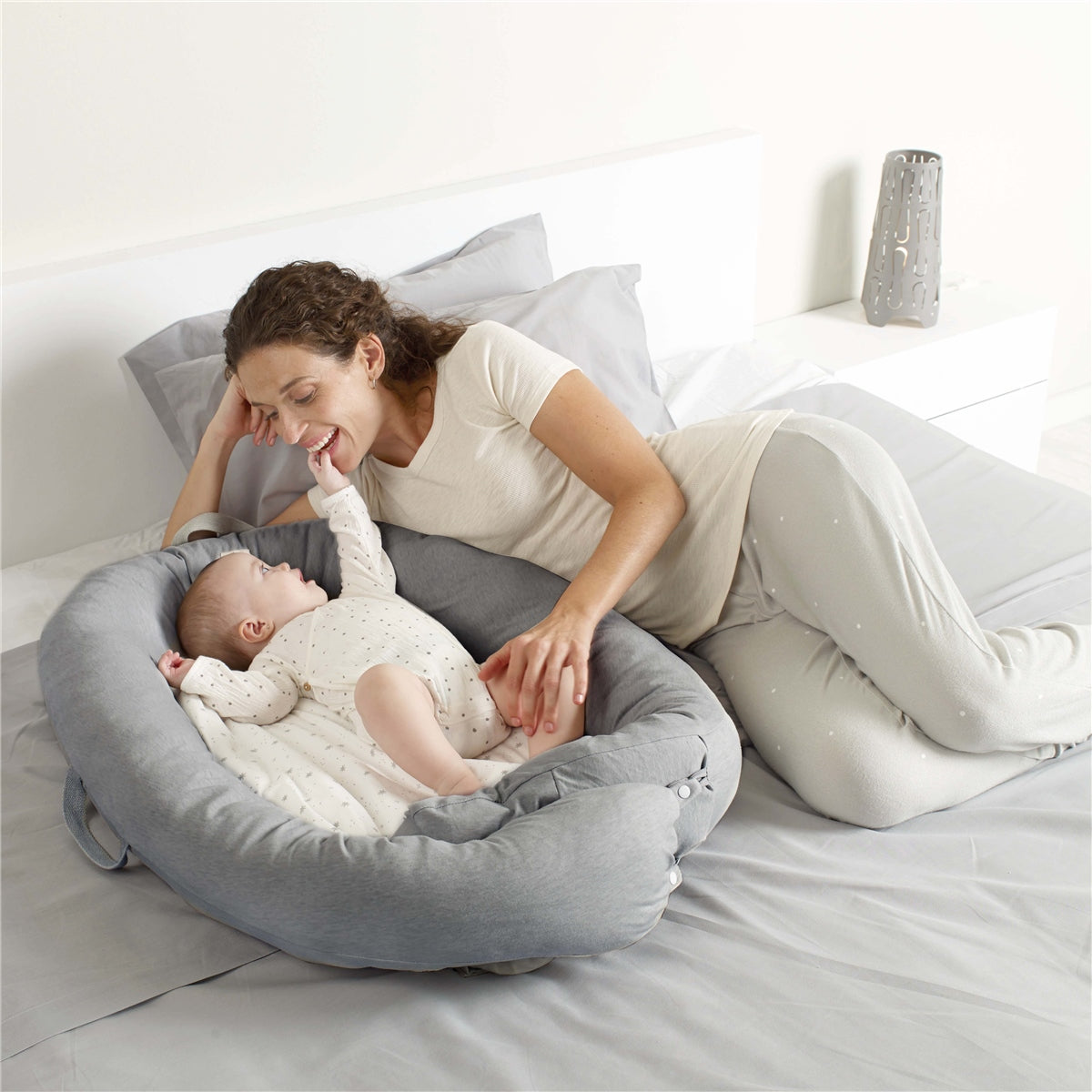 Woman playing with a baby in a gray baby bed on a bed.