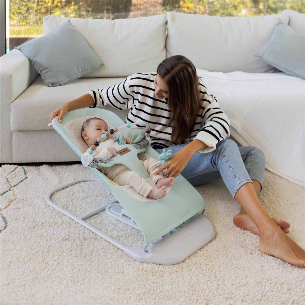Woman sitting on the floor with a baby in a green baby bouncer in a living room.