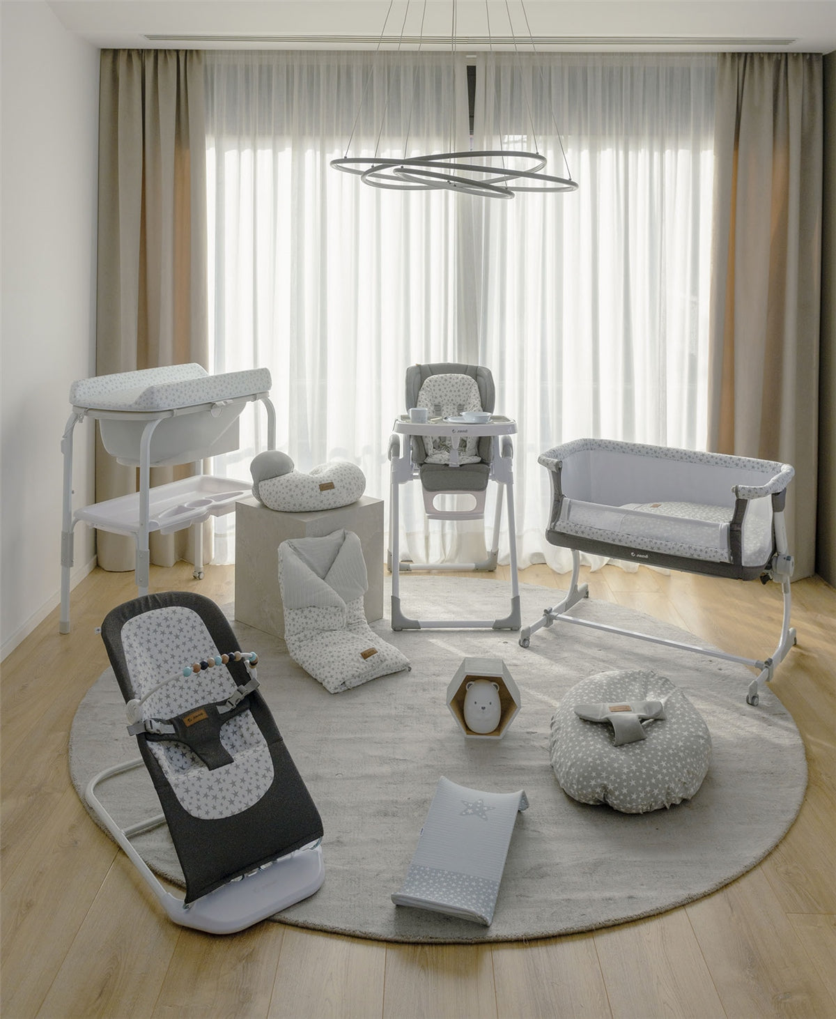 Collection of baby furniture including a high chair, bouncer, and crib in a room with curtains and a rug.