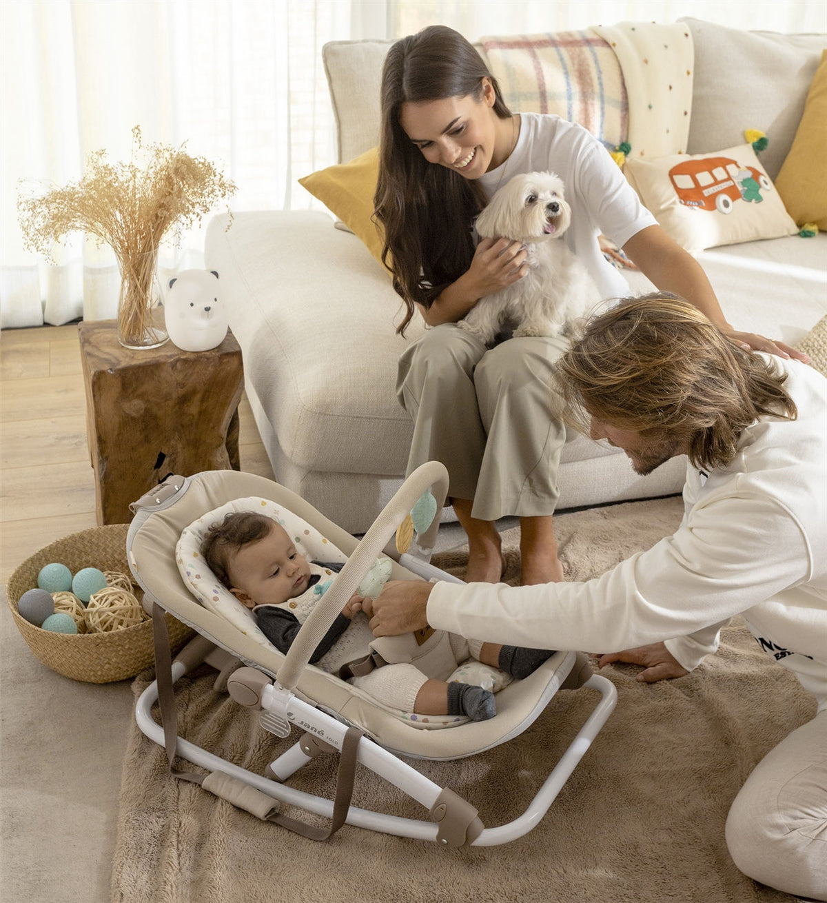 Woman holding a dog, man interacting with a baby in a bouncer, and another baby lying on a couch.