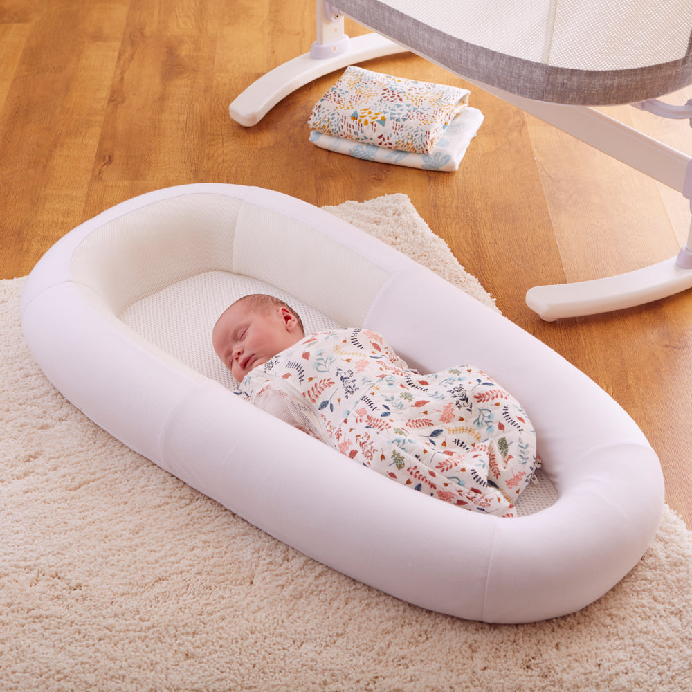 Baby sleeping in a white soft baby bed on a wooden floor with a crib in the background.
