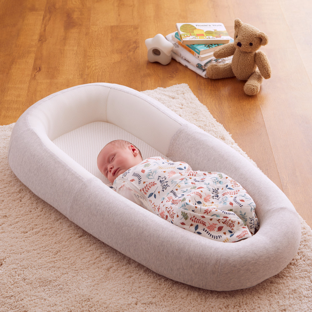 Baby sleeping in a white bassinet on a wooden floor with a teddy bear and books in the background.