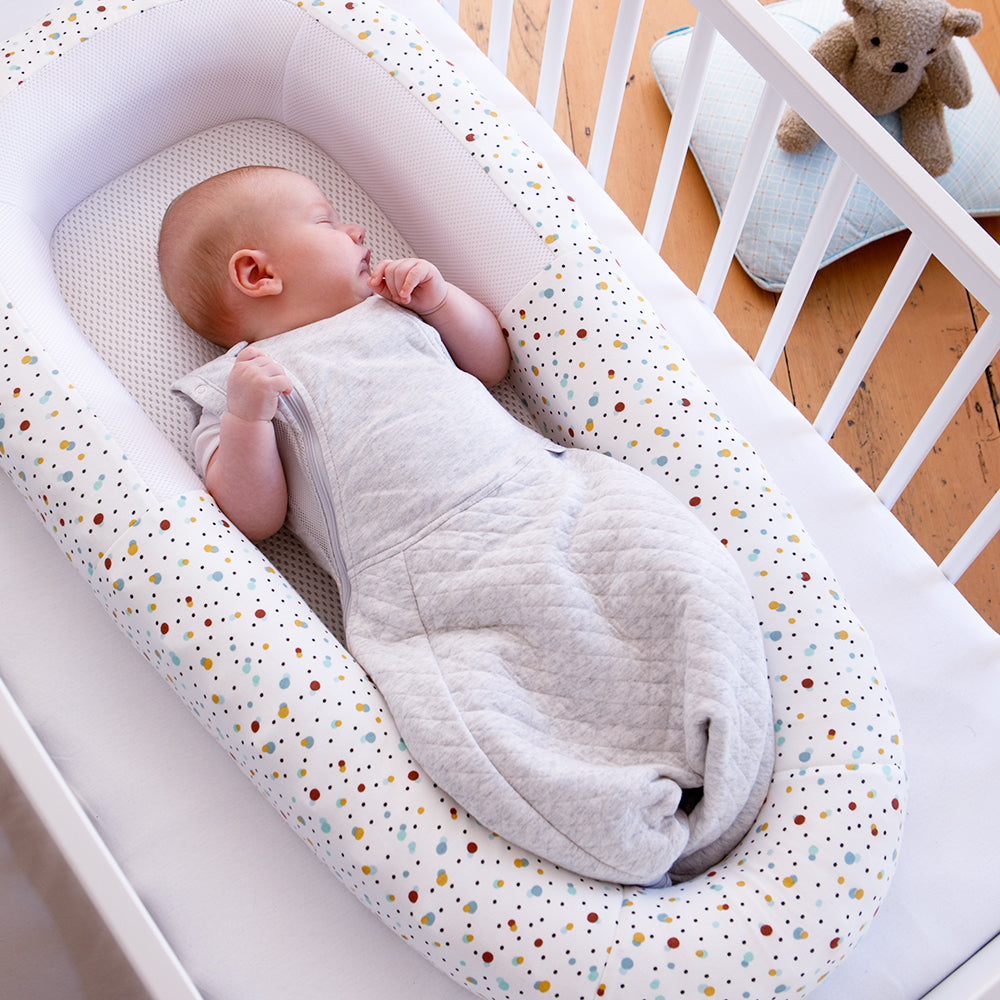 Baby wrapped in a grey swaddle lying in a polka dot baby bed in a crib.