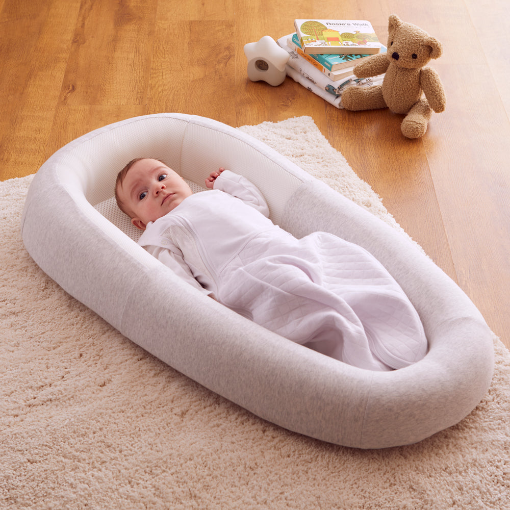 Baby lying in a white and grey baby bed on a wooden floor with books and a teddy bear.