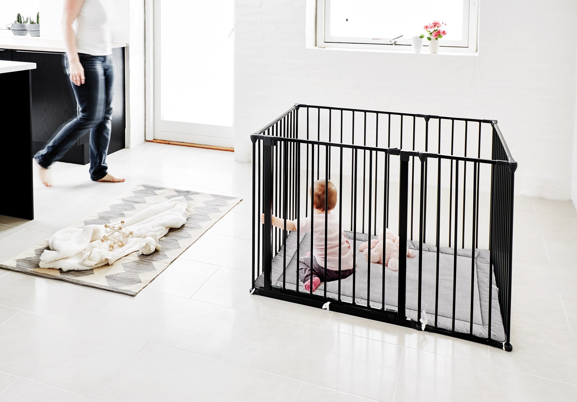 Baby inside a black playpen with a person standing nearby in a room.