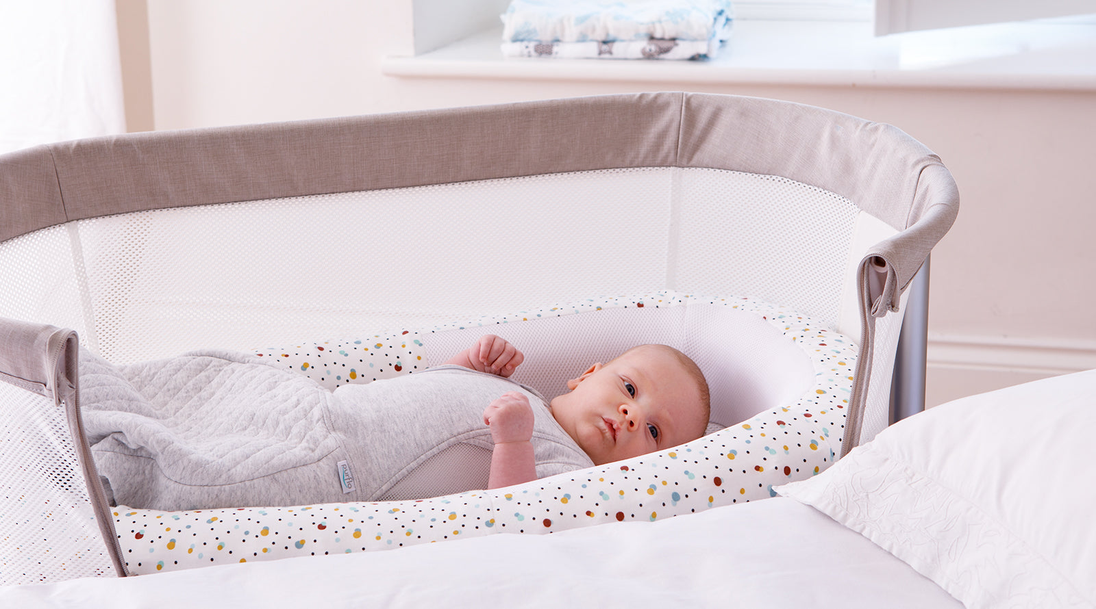 Baby lying in a white crib with a grey rail in a softly lit room.