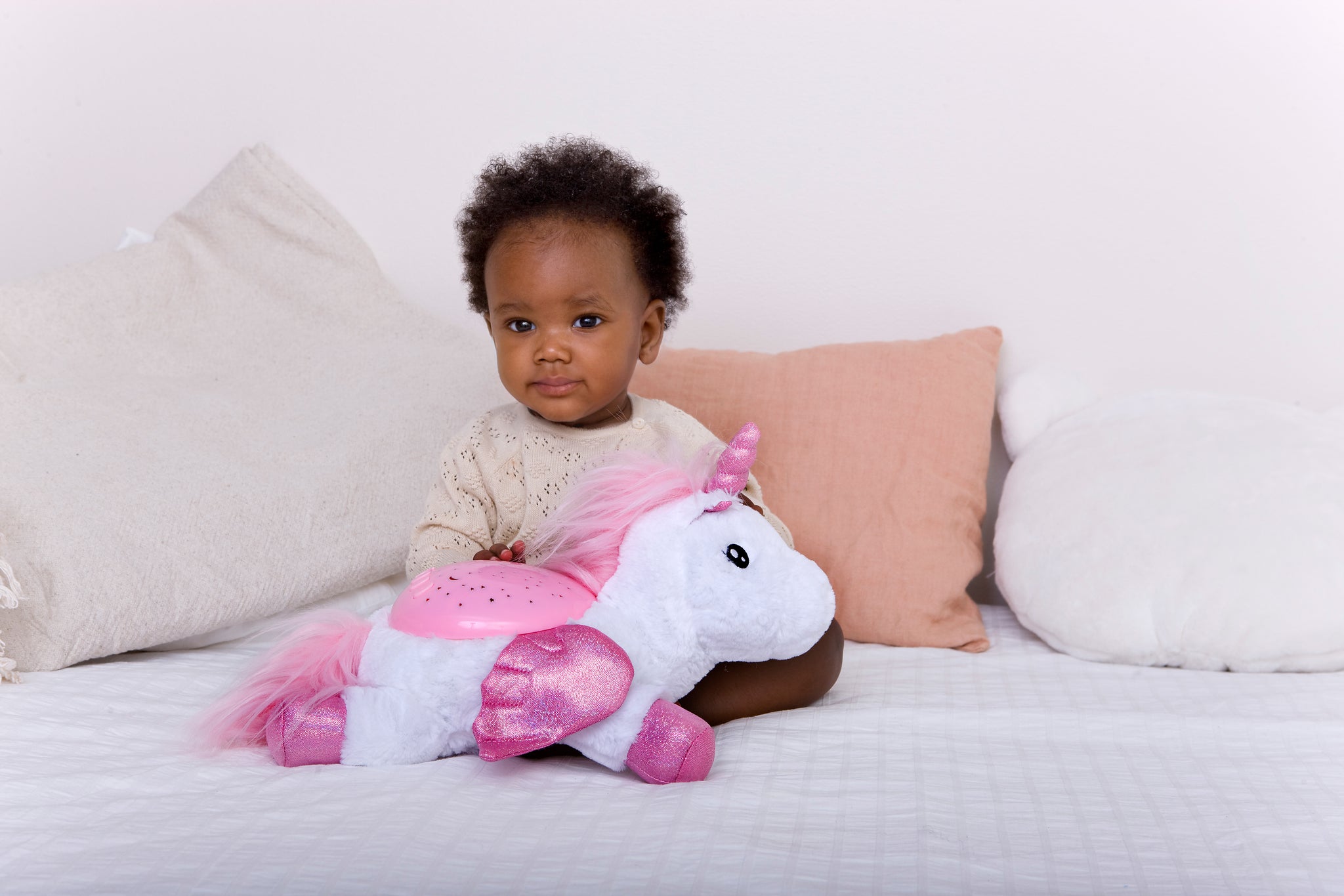 Child holding a pink and white unicorn plush toy on a bed with light-colored pillows.