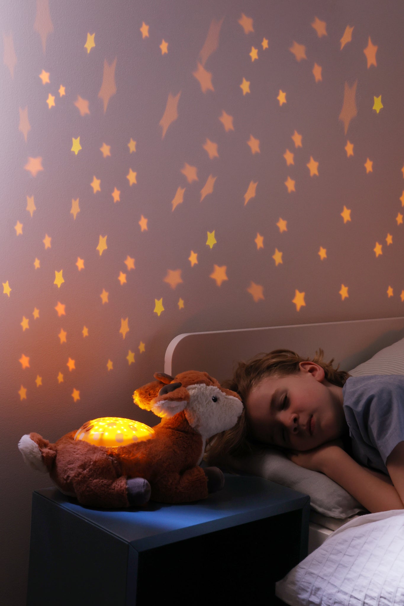 Child lying in bed with a glowing plush toy under a star-lit wall.