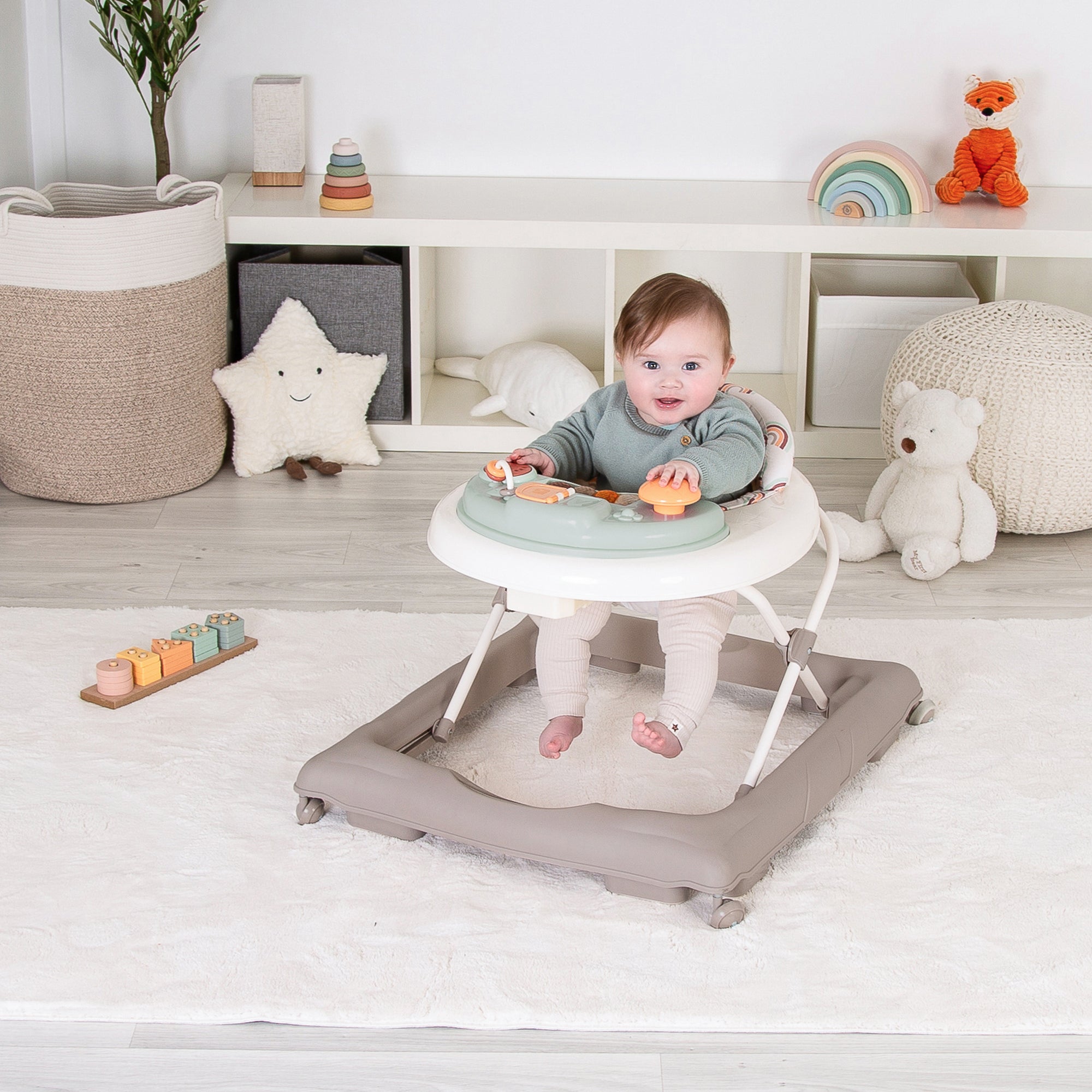 Baby sitting in a white baby walker with a gray mat on a wooden floor, surrounded by toys and decor.
