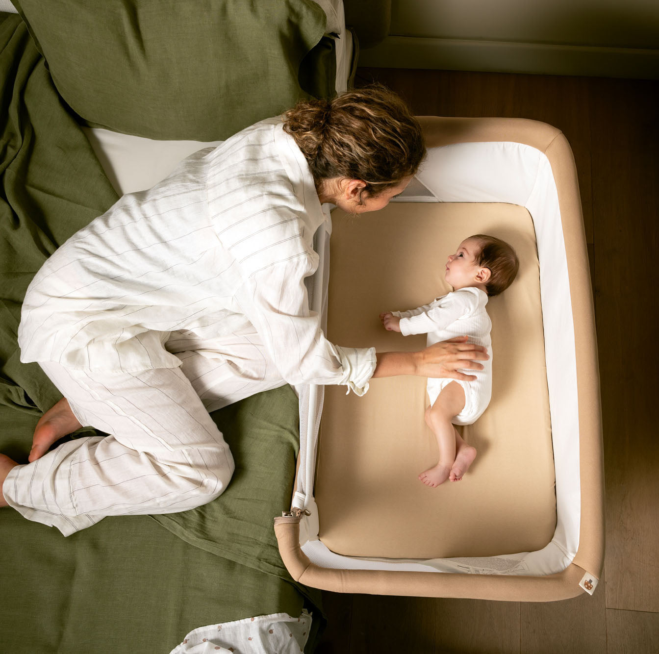 Woman changing a baby in a portable crib next to a bed.
