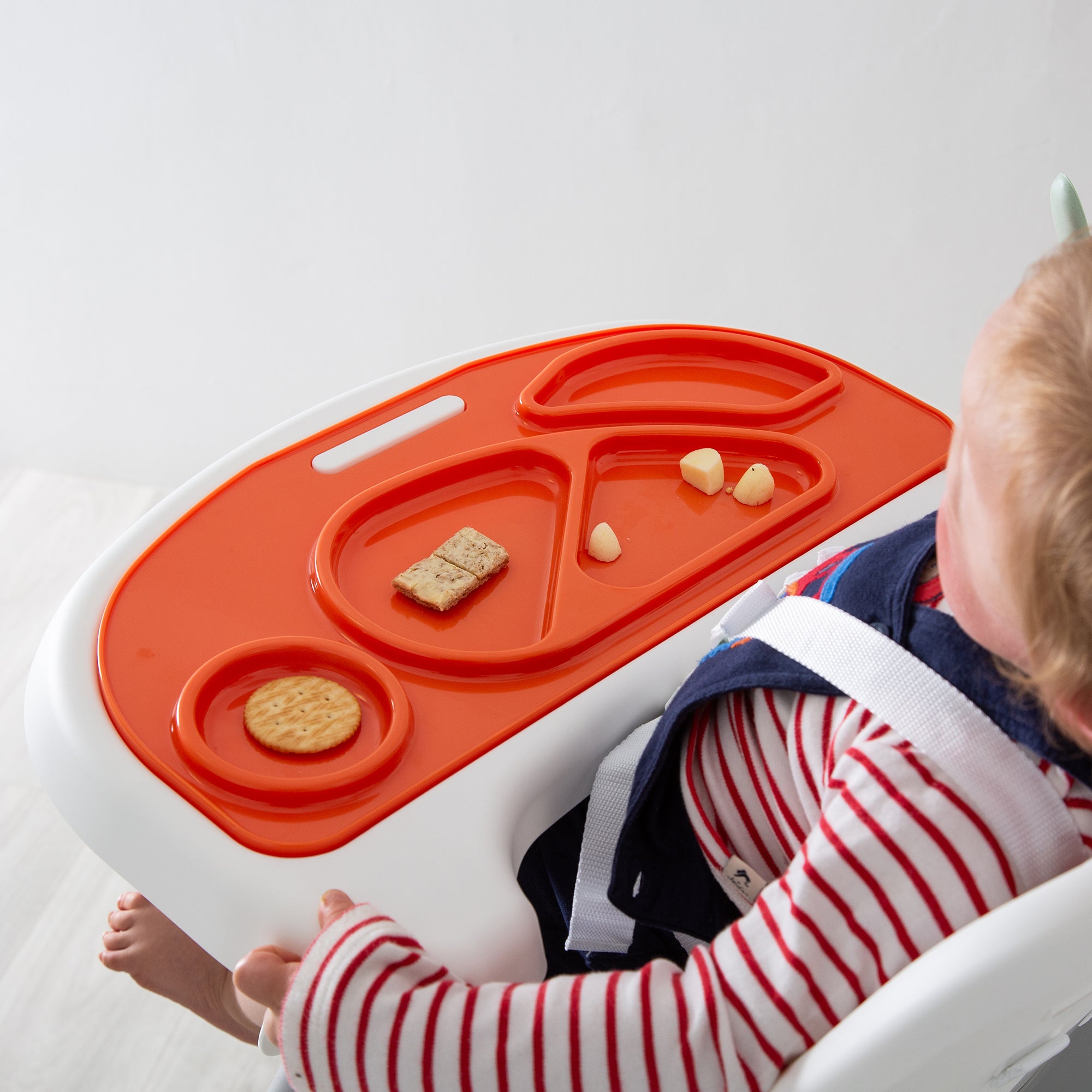 Child sitting on a white chair with a orange and white tray in front of them, containing small food items.