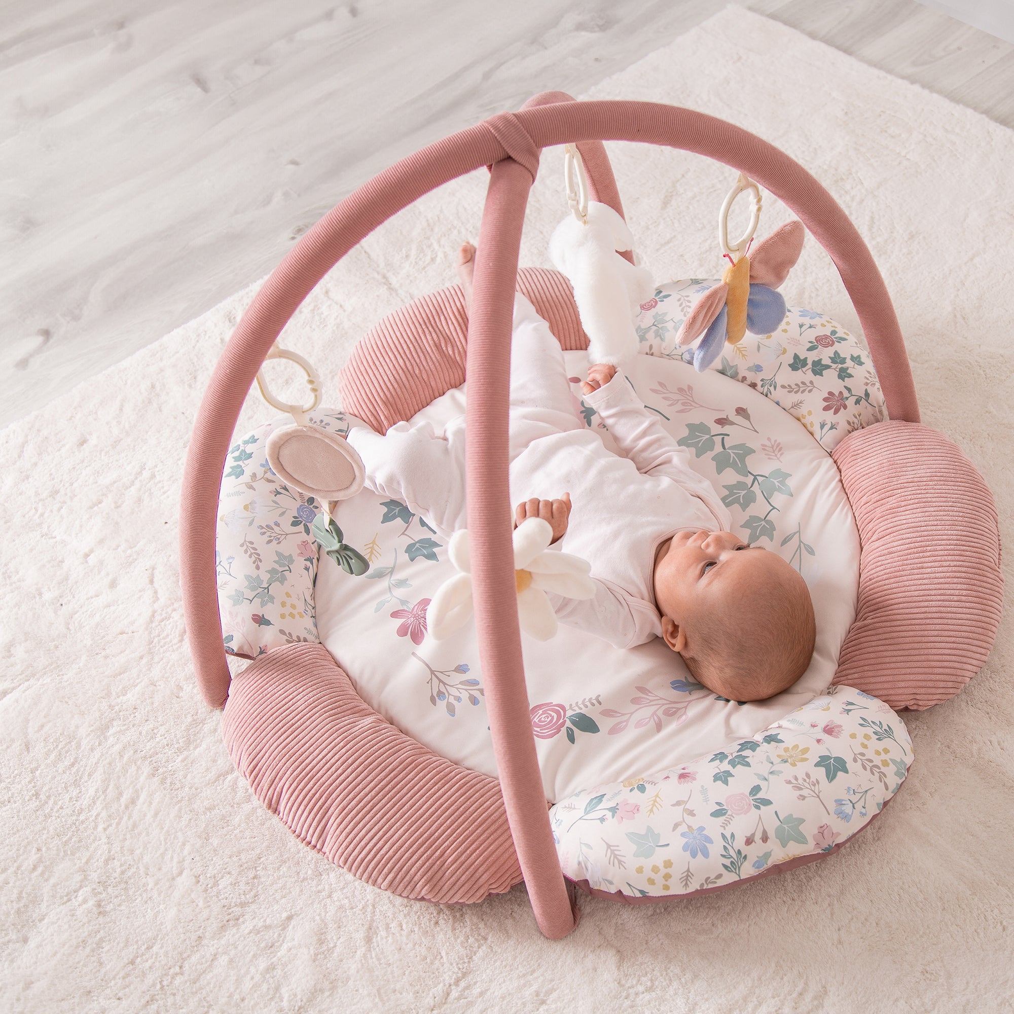 Baby lying in a pink and floral baby playgym on a light wooden floor.