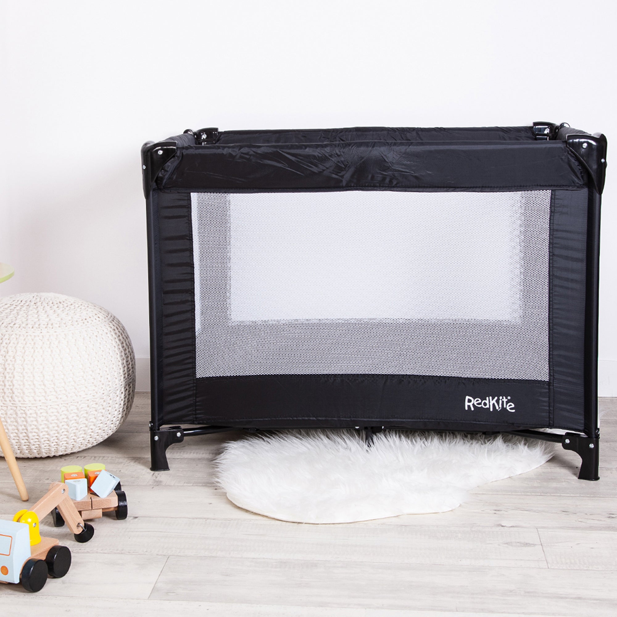 Black baby playpen with mesh door on a light wooden floor, surrounded by toys and a white rug.