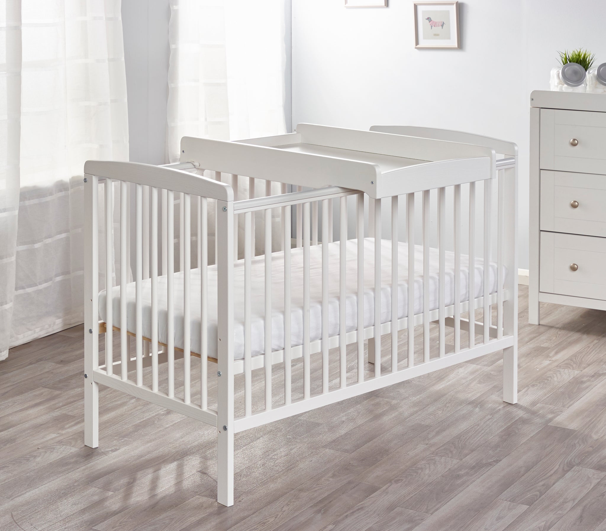 White cot with changing table in a room with a dresser and window.